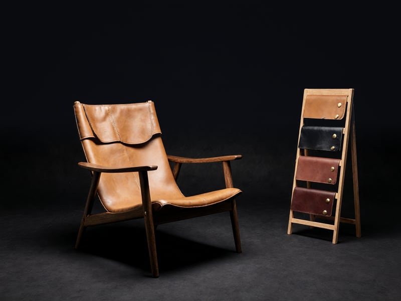 A walnut-framed lounge chair with a tan leather sling, beside an upright wooden rack of leather finish samples.