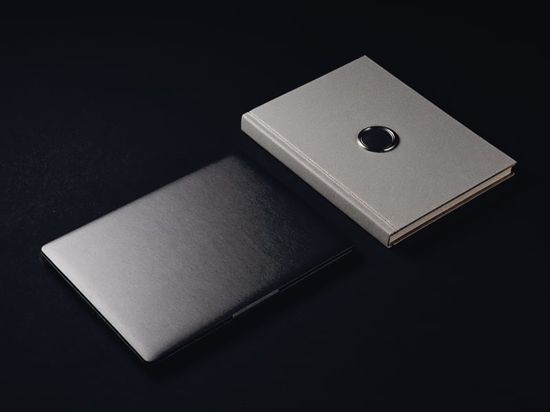 A closed, unbranded laptop lying on a dark stage beside a bound document carrying a blind-embossed seal.